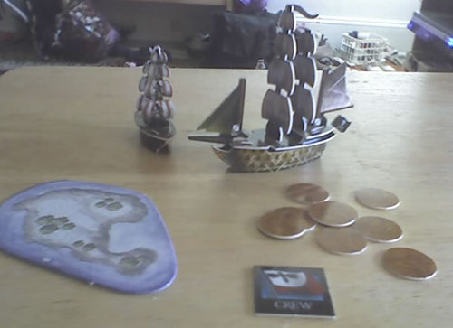 Two small pirate ships, constructed from plastic components, "sail" on a table. An island appears to the left; a number of gold tokens representing treasure appears to the right.