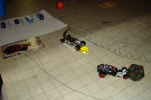 Two small Matchbox cars race across a square grid. Multisided dice indicate initiative; a character sheet can be seen to the left.