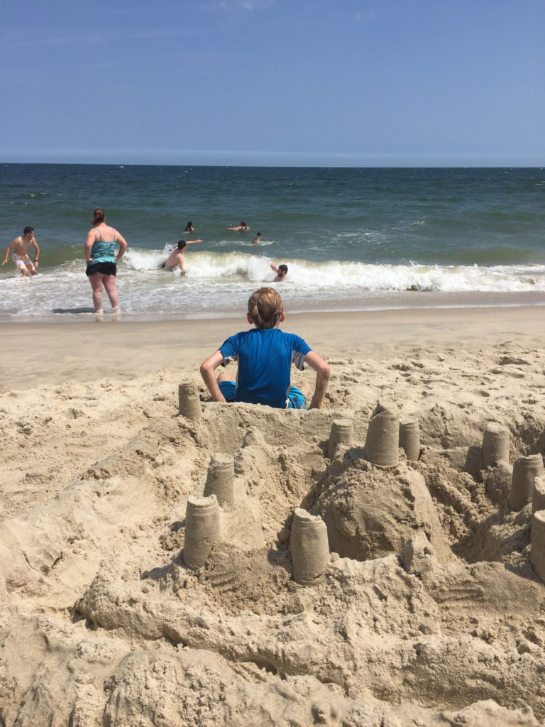 A sand castle fills the foreground while a 10-year-old boy sits just in front of it. The steel-blue ocean is in the background.