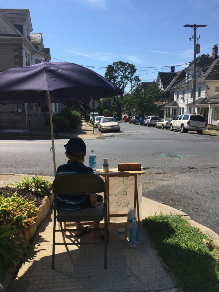 A young boy sits under an umbrella while he sells lemonade at a stand. A city street is in the background.