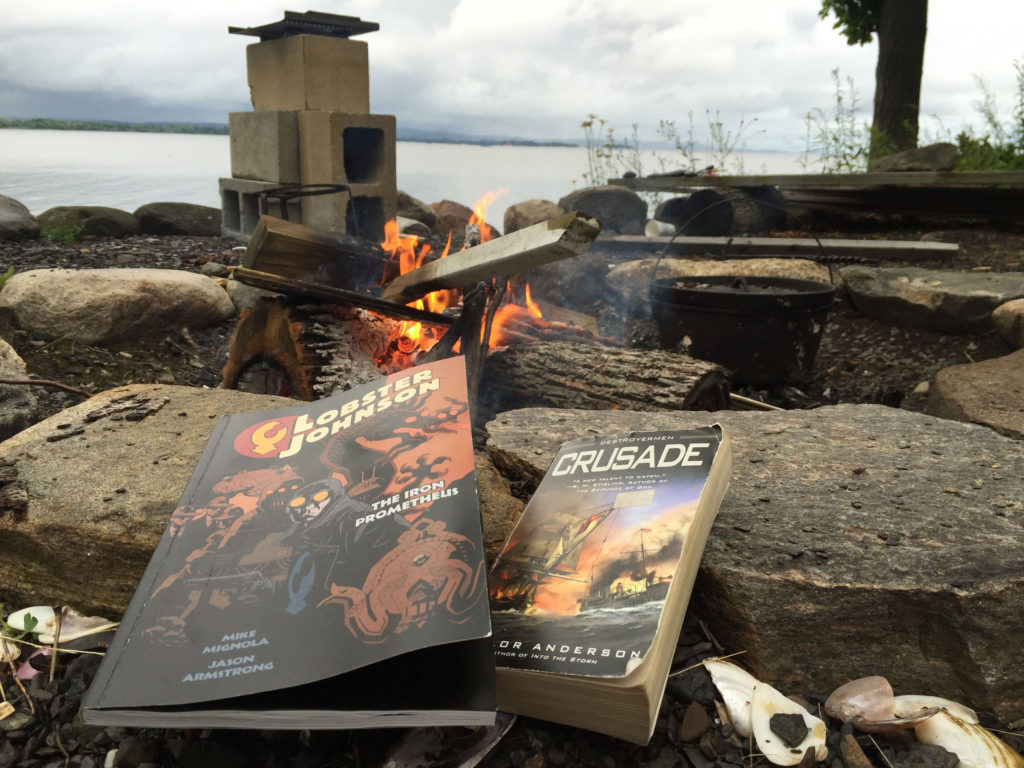 Two books -- Lobster Johnson and Crusade -- rest on rocks next to a fire pit. A fire can be seen burning in the upper portion of the photograph. The sky in the background is overcast.