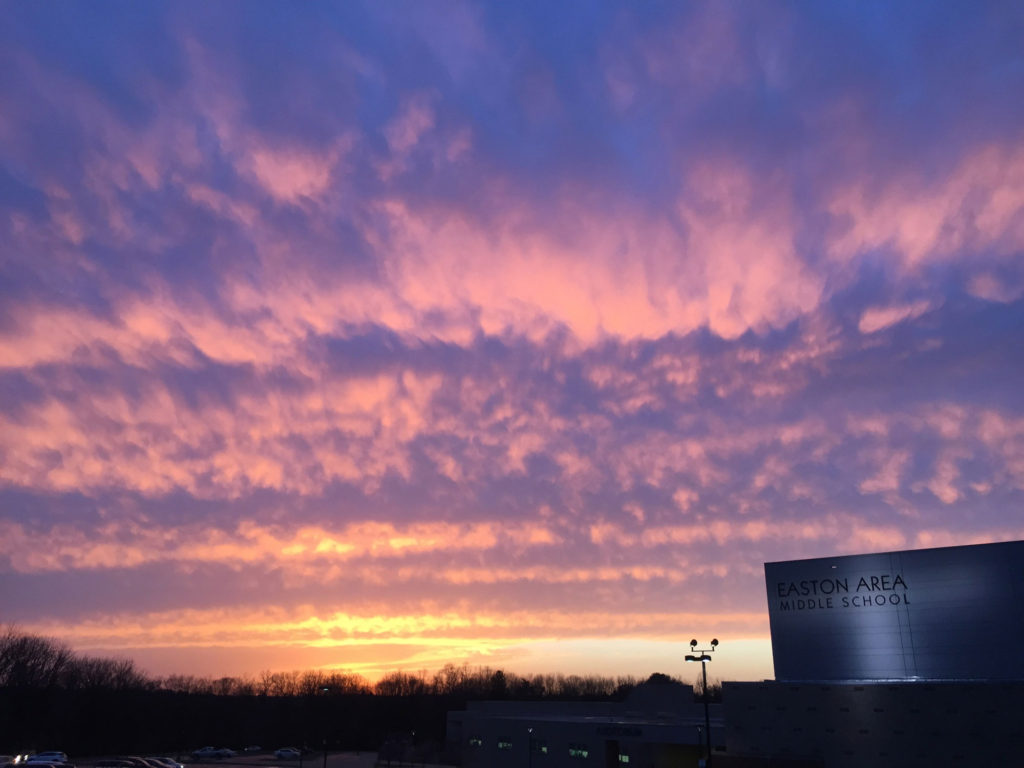 A yellow, pink, and purple sunset dominates this picture. In the lower righthand corner is a block-ish school illuminated by spotlights.