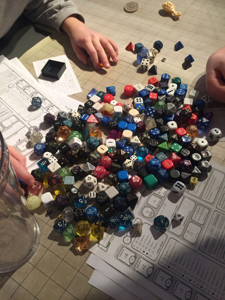 A huge pile of dice fills the picture, overflowing onto character sheets. Boys' hands reach in select dice to use in a game.