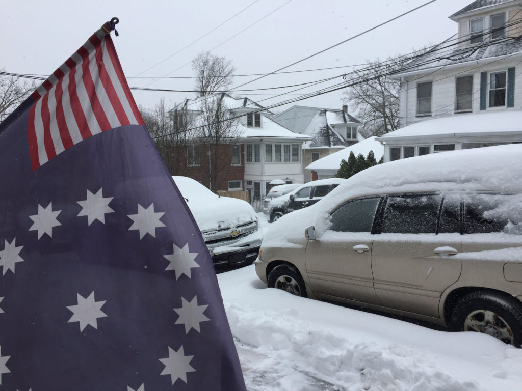 An blue flag with white stars is in the foreground, while snow covered cars appear in the background.