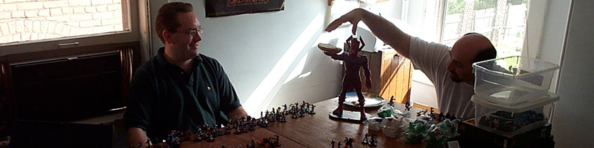 Two men sit at a table. Small plastic HeroClix figures stand arrayed on a large wooden table while a much larger comic book figure (Galactus) holds a bagel.