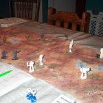 Grey and white robot-like figurines battle on a hexagonal map. The lower left corner of the photo shows a rulebook and some dice.