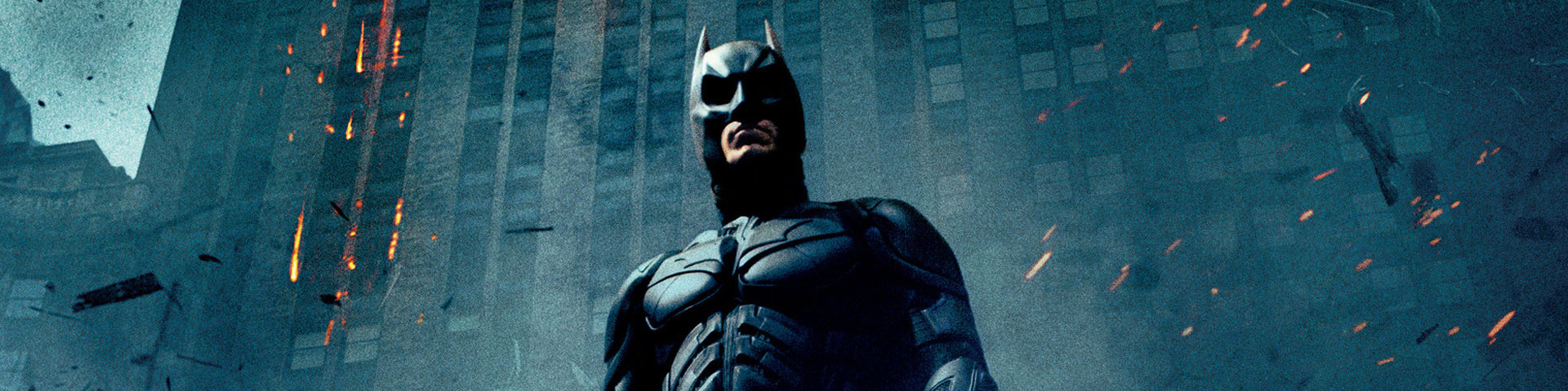 Batman, in his bat armor, stands in front of an office building. Smoke, ash, and cinders swirl through the air.