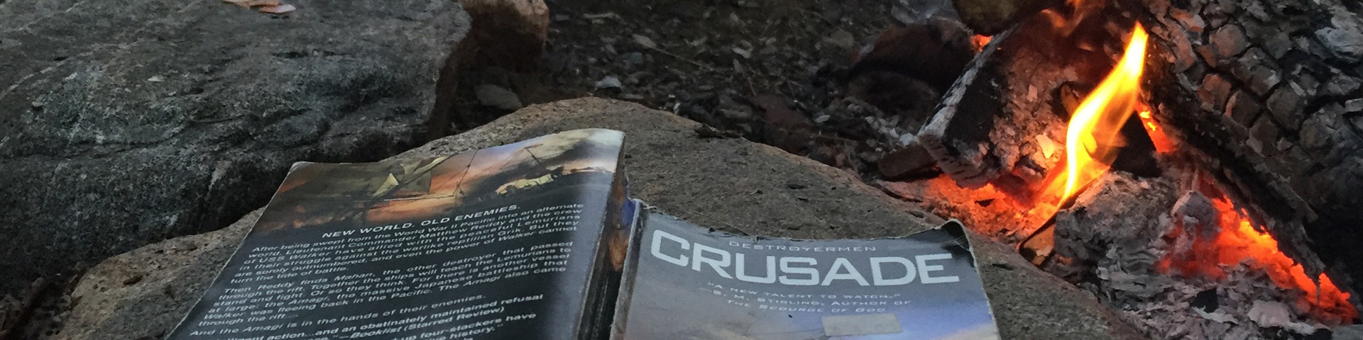 The paperback book "Crusade" lies face down on a rock next to a fire.