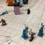Heroes battle monsters in the foreground, while elemental figurines (representing water, fire, and earth) appear in the distance.
