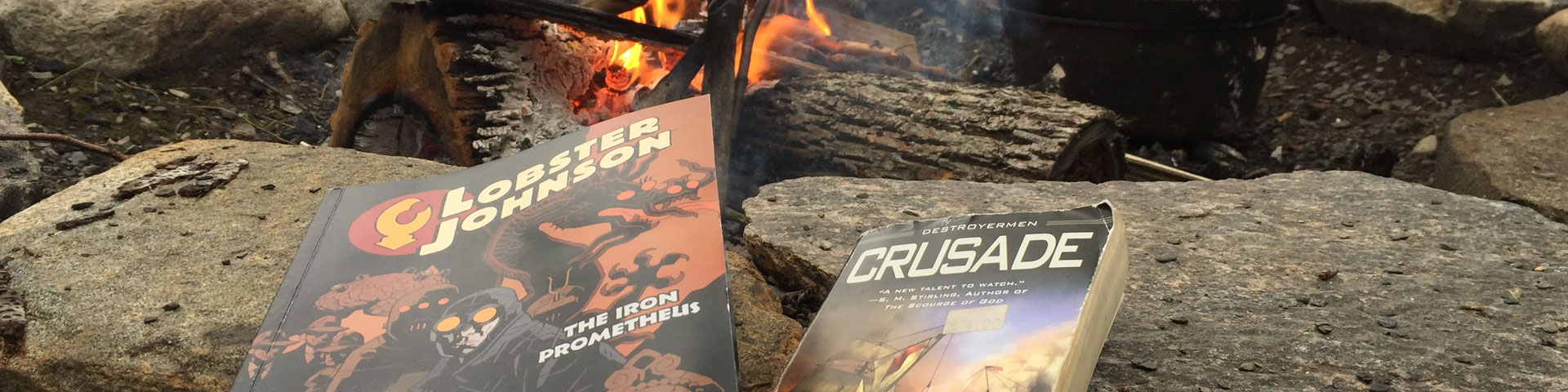 Two books -- Lobster Johnson and Crusade -- rest on rocks next to a fire pit. A fire can be seen burning in the upper portion of the photograph.