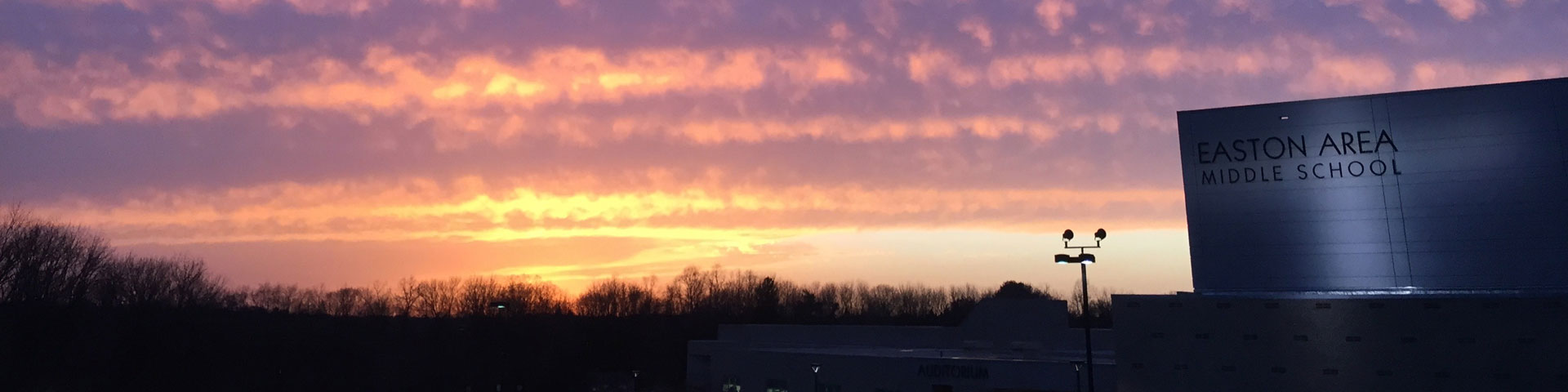 A yellow, pink, and purple sunset dominates this picture. In the lower righthand corner is a block-ish school illuminated by spotlights.