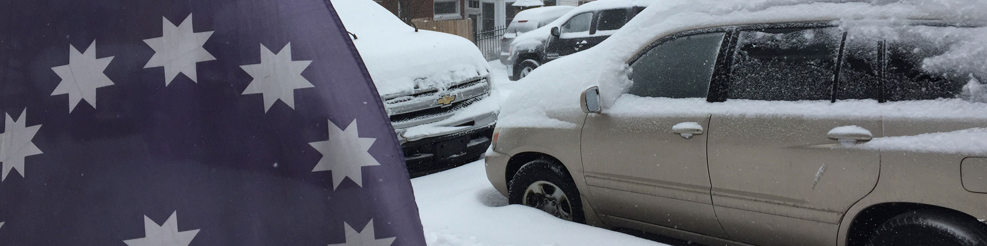 An blue flag with white stars is in the foreground, while snow covered cars appear in the background.