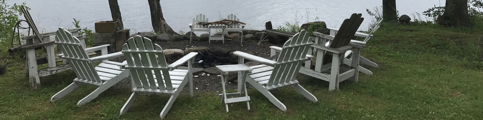 Adirondack chairs gathered around a firepit near a lake.