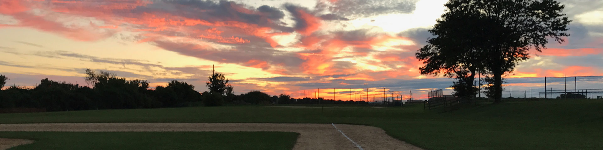A beautiful sunset filled with tinges of orange, red, and yellow. A baseball field appears in the foreground; a large tree is silhouetted off to the right in the background