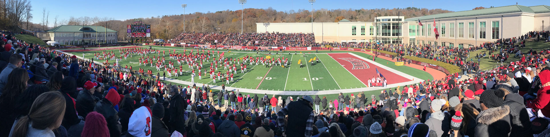 A panoramic view of a football stadium packed with people. A band in red, black, and white uniforms performs on the field.