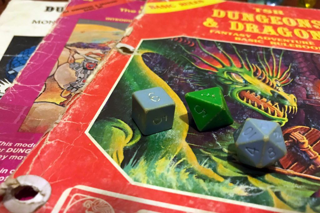 Three old dice - two blue, one green - are laying on top of several battered role-playing game supplements from the 1980s.