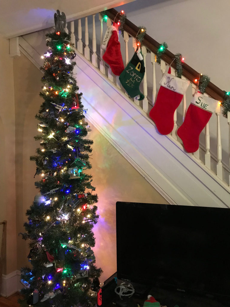 A tall Christmas tree rises next to a set of stairs decked out with stockings. A TV appears to the right.