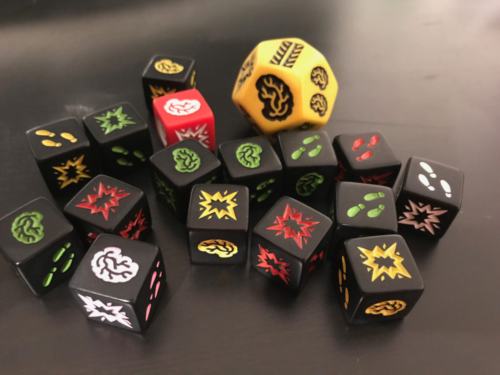 Zombie-themed dice on a black tabletop. The dice have three types of faces: brains, shotgun, and feet. There's also large yellow d12 representing a school bus.
