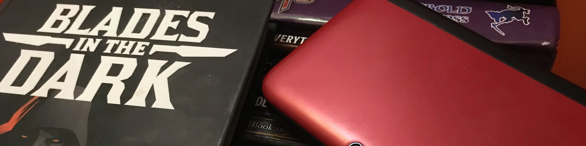 A role-playing game book lays next to a red Nintendo 3DS portable game console. Other RPG source books can be seen beneath the objects.