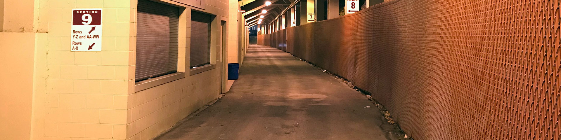 The passageway underneath the stadium seating. A chainlink fence is on the right; closed and shuttered food service areas are on the left.