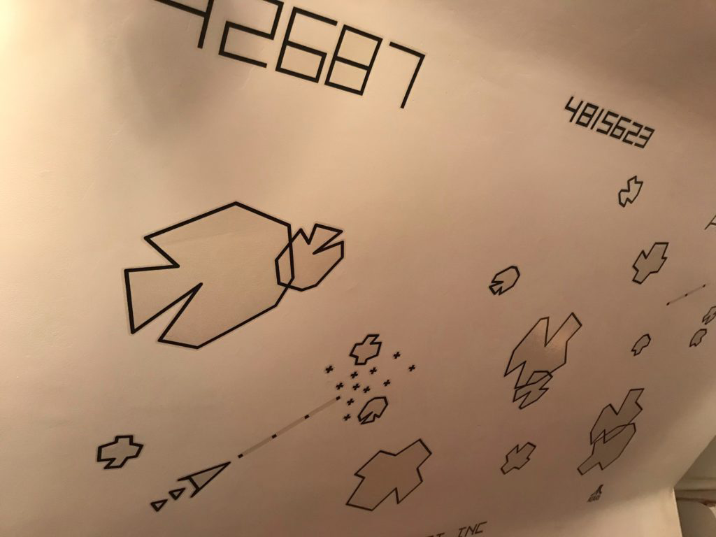 Black and white decals of vector-drawn asteroids and a space ship on a white wall. The space ship is blasting apart an asteroid. The score and high score can be seen at the top of the photo.