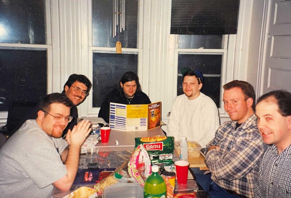 A group of gamers gathered around a dining room table. A D&D 2nd Edition DM screen can be seen at the head of the table.