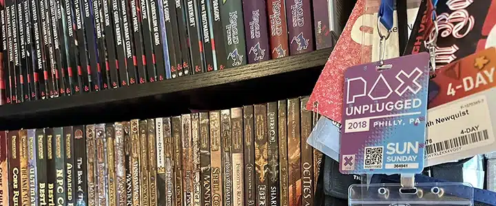 A collection of role-playing game books on a bookshelf, some of which are featured in RPG a Day 2023 posts. A bunch of badges from game conventions fills the right side.