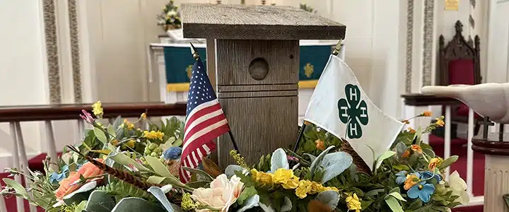 A bluebird box surrounded by a floral wreath. Two flags - an American flag and a 4-H flag - flank the birdhouse.