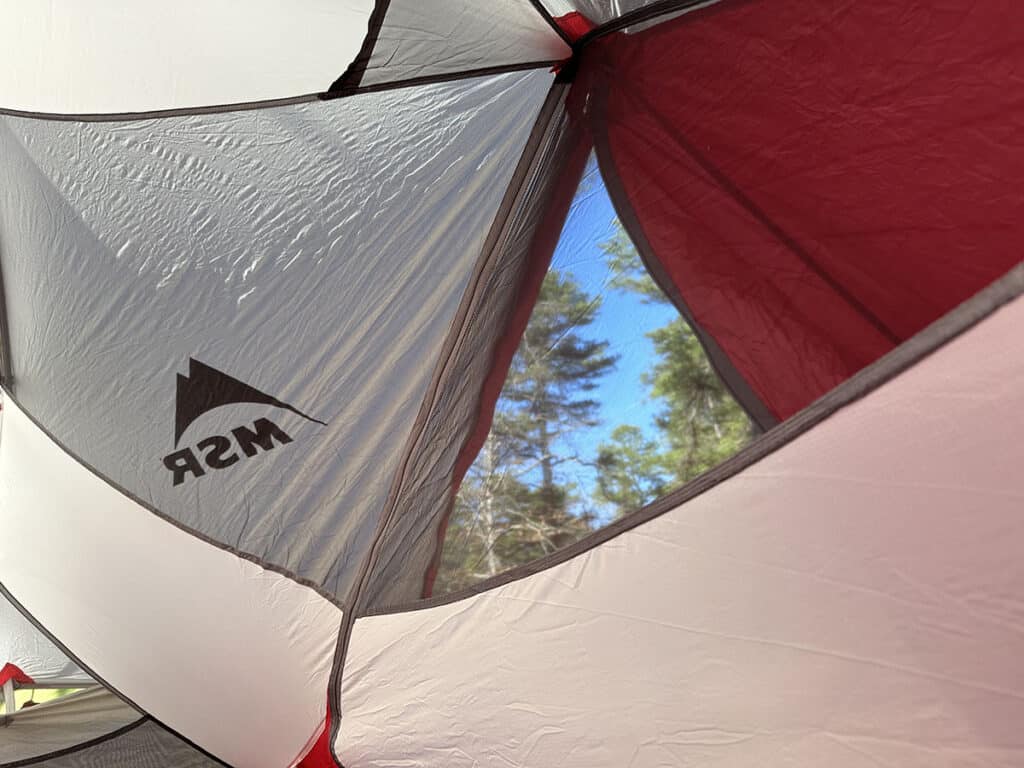 A view of the outside from inside of a tent. Red and white panels form the perimeter of the view; a triangular opening reveals pine trees and blue skies