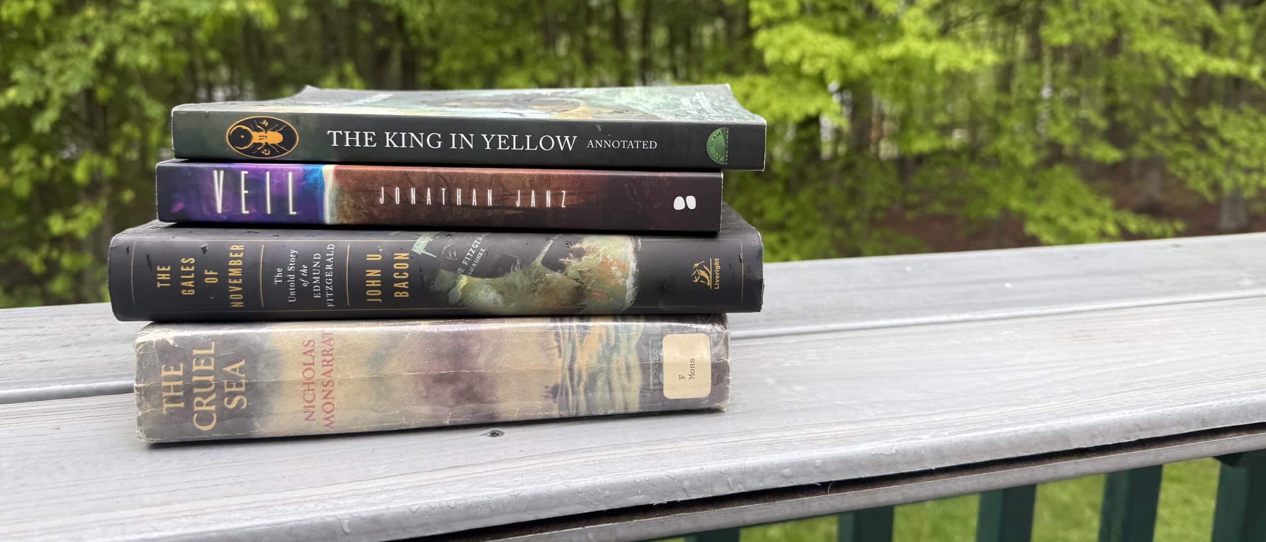 Four books stacked on a flat grey table. Green trees appear in the backgrond.